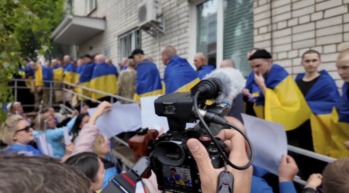 Camera operator filming Ukrainian veterans wrapped in Ukrainian flags outside a building